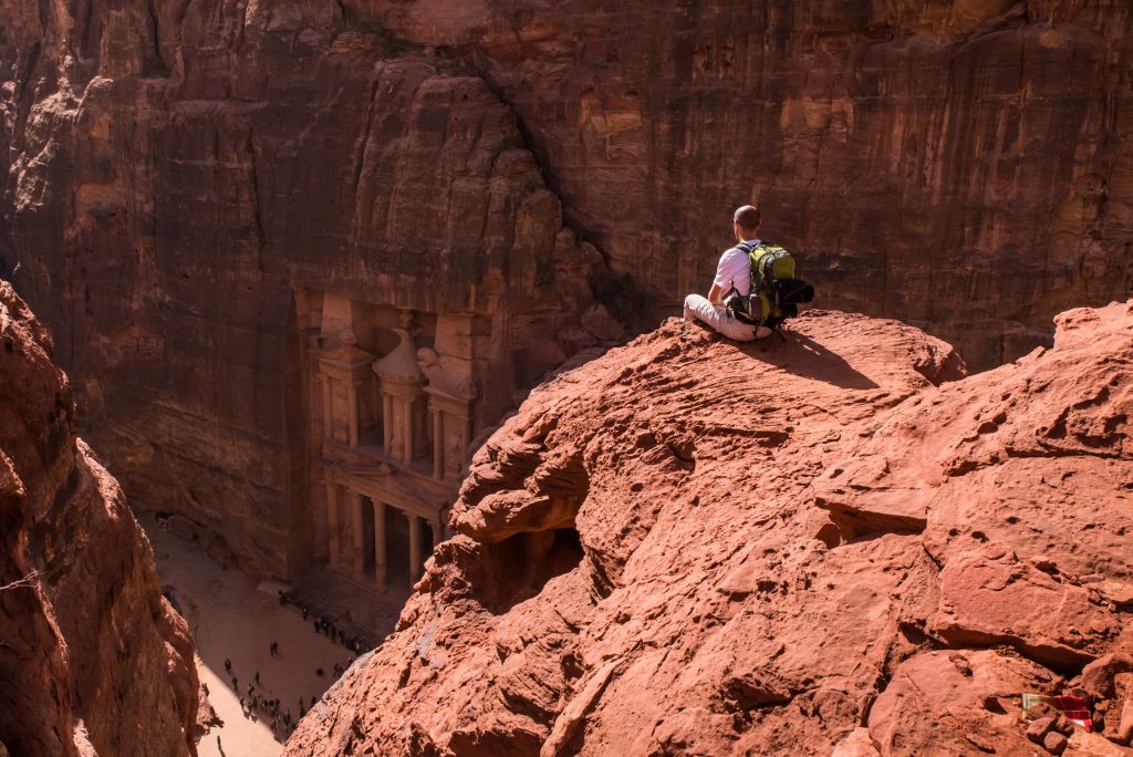 Solo traveler with backpack seated on a cliff over the Treasury, Petra, Jordan