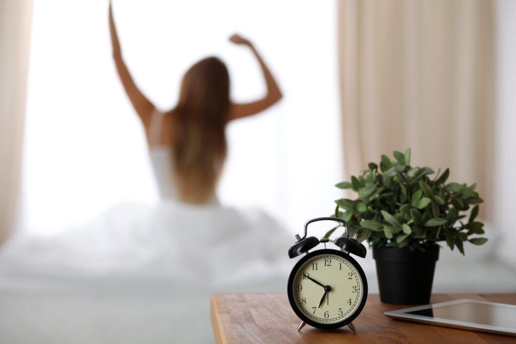 The alarm clock on the nightstand has already gone off early in the morning to wake up the woman who is stretching out in bed in the background.  Waking up early, not getting enough sleep, oversleeping concept.