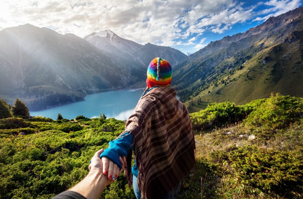 Tourist woman in rainbow hat and brown poncho holding man by hand and going to the lake in the mountains