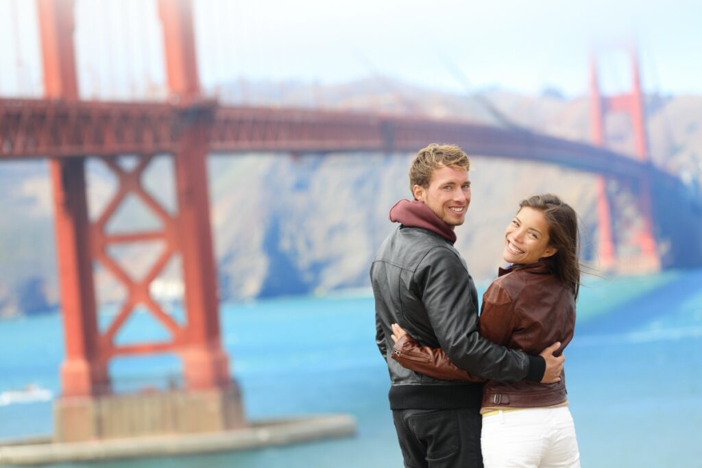 Golden gate bridge happy travel couple in San Francisco, USA smiling at camera. Young interracial hipster couple enjoying the view at the famous travel landmark.
