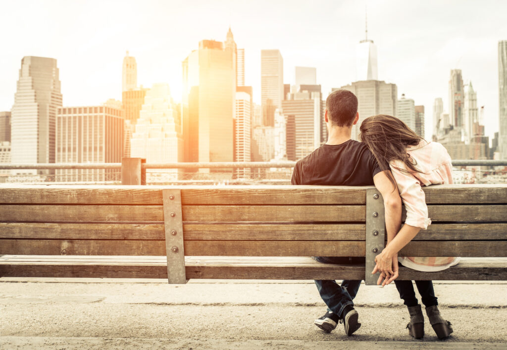 couple relaxing on New york bench in front of the skyline at sunset time having a safe travel experience