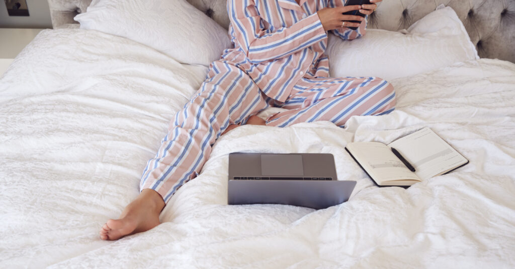 Close Up Of Businesswoman In Pyjamas Sitting On Bed With Laptop And Mobile Phone Working From Home