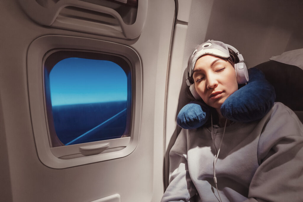 An Asian girl sleeps in her seat on the plane near the window, wearing a mask and a sleeping pillow.  The concept of traveling with comfort and jet lag