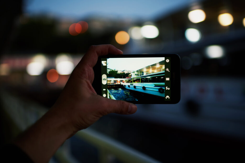 Person taking a photo of truistic sail boat using smart phone camera, male hand holding cell phone while taking a photograph of night lights city in travel,taking a picture of outdoors,blur background