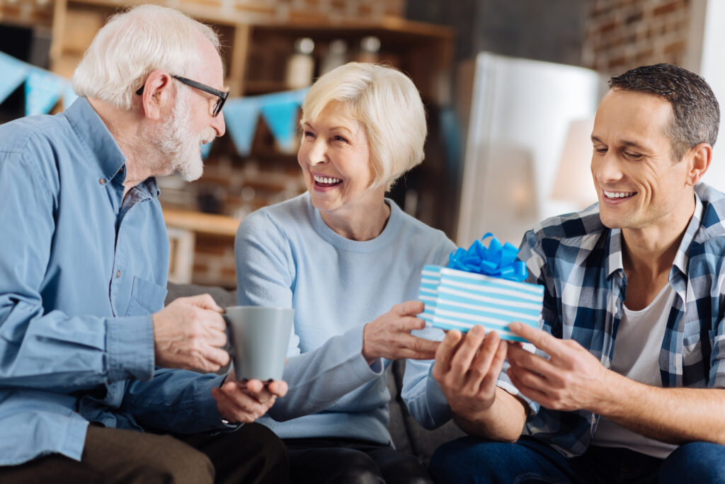 happy birthday.  An optimistic young man sits on the sofa next to his elderly parents and gives a birthday gift to his mother while sharing happiness with her husband
